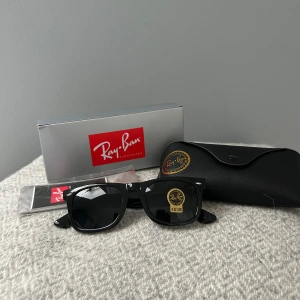 Svarta Ray-Ban Wayfarer solglasögon - Ray-Ban Wayfarer solglasögon med klassisk svart båge i plast och mörka linser. Modellen är RB2140 med stilren och tidlös design. Säljes med originalfodral och box. Perfekt för dig som vill ha en ikonisk look och skydda ögonen mot solen.