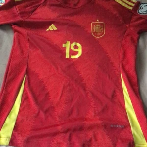 Spanien Lamine Yamal #19 fotbollströja - Säljer en officiell Spanien landslagströja från Adidas med Lamine Yamal och nummer 19 i gult på ryggen. Tröjan är röd med gula detaljer och har korta ärmar, samt tryckta landslagsmärken och Aeroready-material för extra komfort. Perfekt för dig som älskar fotboll och vill ha en snygg matchtröja., skriv vid intresse.