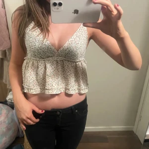 Blommig croppad linnetopp med volang - Supersöt croppad linnetopp i ljusbeige med små blommor i rosa och grönt. Toppen har v-ringad hals, breda axelband och en volang nedtill som ger en flowy vibe. Perfekt för varma dagar och chill häng. Materialet känns lätt och luftigt.