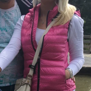 Rosa quiltad dunväst med huva - Snygg rosa dunväst med quiltad design och dragkedja framtill. Västen har en praktisk huva med knappar och svarta detaljer längs kanterna. Perfekt att slänga över en hoodie eller långärmad tröja för extra värme. Fickor med dragkedja på sidorna.