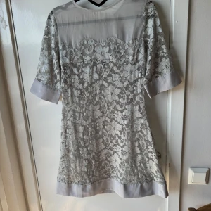 Grå spetsklänning från Repeat - Snygg grå kortklänning från Repeat med halvärmar och blommigt spetsmönster. Klänningen har transparent parti upptill, breda manschetter och dragkedja i ryggen. Perfekt för dig som gillar romantisk stil och detaljer som spets och blommor.