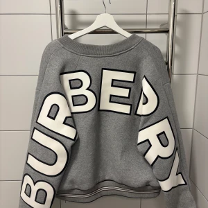 Grå Burberry bomberjacka med logga - Säljer en grå bomberjacka från Burberry med stor vit logga på ryggen och ärmarna. Jackan har ribbade muddar, två fickor med knapp och dragkedja framtill. Tillverkad i mjuk bomullsmix, perfekt för dig som gillar streetwear och snygga detaljer. Storlek M