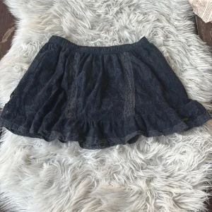 Svart spetskjol Abercrombie & Fitch - Supersöt svart kortkjol från Abercrombie & Fitch med volangkant och vackert spetsmönster. Kjolen har elastisk midja och är fodrad för extra komfort. Perfekt för dig som gillar romantisk och feminin stil.