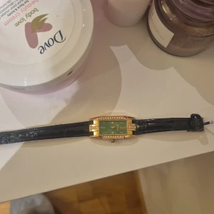Grön och guldig klocka med strass - Snygg rektangulär klocka från Delia med grön urtavla, guldiga detaljer och strass runt boetten. Svart, glansigt armband med klassiskt spänne. Perfekt för dig som gillar att sticka ut med accessoarer som har lite lyxig vibe. Fick den som present, pris kan diskuteras om ni vill.