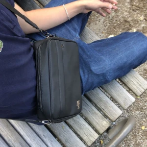 Svart axelväska från Lacoste - Snygg svart axelväska från Lacoste med klassisk krokodillogga i metall på framsidan. Väskan har en rektangulär form, justerbar axelrem och ett stort huvudfack med dragkedja. Materialet är slitstark syntet med diskret textur. Perfekt för att bära dina essentials.