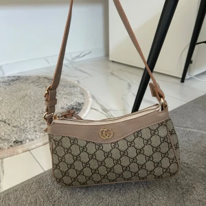 Gucciväska  - Snygg beige axelväska med klassiskt GG-mönster i brunt och detaljer i ljusbrunt skinn. Väskan har guldfärgad GG-logga framtill och dragkedja upptill. Justerbar axelrem med guldfärgade metalldetaljer. Perfekt storlek för det viktigaste.