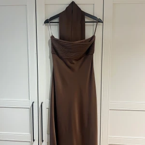 Brun axelbandslös långklänning - Säljer en brun satinklänning med scarf från australiensiska Runaway the Label. Den är använd en gång och är gjord i tjockt, lyxigt polyester-material med silikonband i bysten så att den sitter på plats. Längden slutar precis ovanför fötterna, jag är 170 cm. Nypris var cirka 2900 kr inklusive frakt, tull och moms. Säljer även samma modell i svart i en annan annons. För fler bilder är det bara och höra av sig.