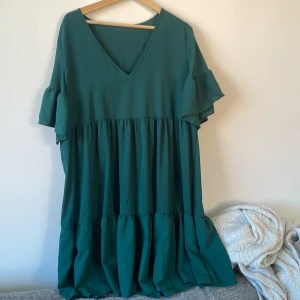 Grön volangklänning från Greathe Curve - Säljer en snygg grön klänning från Greathe Curve i storlek OXL. Klänningen har v-ringning, korta volangärmar och flera volanger nedtill som ger en luftig och cool look. Materialet känns lätt och skönt mot huden, perfekt för dig som gillar avslappnad stil.