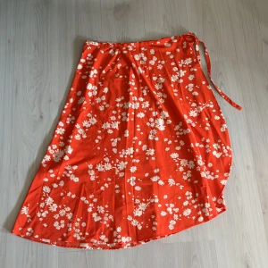Röd blommig omlottkjol från Lindex - Superfin röd omlottkjol från Lindex med vita blommor och knytband i midjan. Kjolen är lång och har en flowy känsla, perfekt för varma dagar. Materialet känns lätt och svalt, och mönstret är riktigt somrigt.