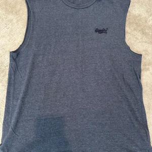 Mörkblå ärmlös linnetopp Superdry - ✨ Superdry Linnetopp ✨ Snygg mörkblå linnetopp från Superdry 🔵 med diskret logga på bröstet 👕. Ärmlös design med rund halsringning för en klassisk och avslappnad stil. 	•	🧵 Tillverkad i mjuk bomull som känns skön mot huden 	•	🌞 Perfekt för varma sommardagar eller lager på lager 	•	👌 Enkel, clean och alltid lätt att matcha  Ett mångsidigt basplagg för en stilren och bekväm look.