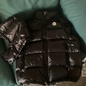 Svart dunväst från Moncler (passar storlek L) - Säljer en svart, glansig dunväst från Moncler med hög krage och två dragkedjefickor framtill. Västen har ett klassiskt Moncler-märke på bröstet och quiltad design som ger en cool och sportig vibe. Perfekt för lager-på-lager under kyliga dagar.