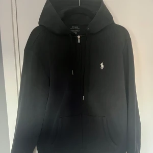 Svart hoodie Polo Ralph Lauren - Svart hoodie med dragkedja från Polo Ralph Lauren. Bra skick men tyvärr en fläck av målarfärg på högerarmen, därav priset. Däremot syns den knappt. Storlek S. Hör av er vid frågor eller funderingar! 