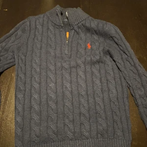 Mörkblå stickad tröja från Polo Ralph Lauren - Säljer en mörkblå stickad tröja från Polo Ralph Lauren med klassiskt kabelstickat mönster och röd logga på bröstet. Tröjan har lång ärm, ribbade muddar och en half zip-dragkedja vid halsen. Perfekt för dig som gillar preppy stil och snygga detaljer.