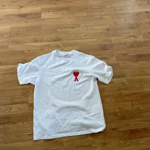 Vit t-shirt från Ami med rött hjärta - Vit t-shirt från Ami Alexandre Mattiussi med ett rött hjärta och bokstaven A broderat på bröstet. Klassisk rund hals och korta ärmar. T-shirten är gjord i mjuk bomull och har en clean. Storlek M