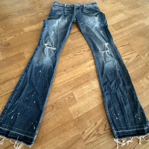 Lågmidjade coola bootcut jeans - Säljer ett par blå lågmidjade bootcut jeans från Guess med slitna detaljer samt vita färgstänk som är en del av designen, de har också  fransade benslut.  Midjemått: 37cm platt,  innerbenslängd: 83cm.   Storlek på etikett; 27, skulle säga att de passar en xs/ ev liten S.           Det är bara att skriva om ni har några funderingar! 💙