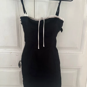 Svart kort klänning med snörning - Snygg svart kort klänning med vita detaljer upptill och snörning framtill. Axelbandslös design med figurnära passform och markerad byst. Perfekt för dig som gillar minimalistisk stil med en twist. Materialet känns mjukt och bekvämt mot huden.