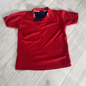 Röd Polo Ralph Lauren t-shirt - Snygg röd t-shirt från Polo Ralph Lauren med klassisk pikékrage och korta ärmar. T-shirten har en marinblå broderad logga på bröstet och är gjord i mjuk bomull. Perfekt för dig som gillar stilrena och sportiga plagg med premiumkänsla.