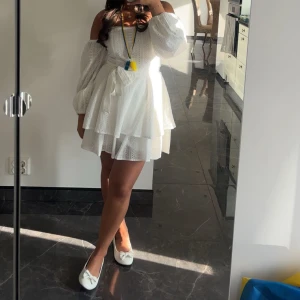 Vit omlott kortklänning med volanger - Supersöt vit kortklänning med omlott och volanger, offshoulder och långa puffärmar. Klänningen har ett knyt i midjan och är gjord i bomull med broderade detaljer. Perfekt för sommarens alla tillfällen, riktigt fräsch och trendig look.