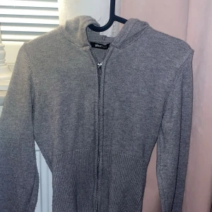 Grå stickad hoodie från Gina Tricot - Säljer en grå stickad hoodie från Gina Tricot med dragkedja framtill och huva. Tröjan har långa ärmar och ribbad nederdel som ger en snygg passform. Perfekt för dig som gillar enkel och stilren look med lite cozy vibes.