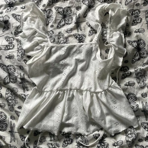 Vit volangblus med knyt och broderi - Superfin vit blus med volangärmar och broderade detaljer. Toppen har omlottkänsla med knyt i ryggen och är sydd i luftig bomull. Perfekt för varma dagar med sin ärmlösa design och romantiska look.