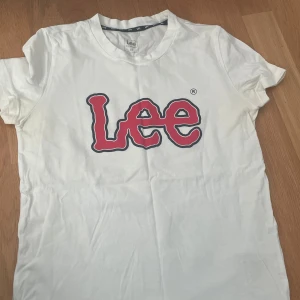 Vit Lee t-shirt med röd logga - Snygg vit t-shirt från Lee med stor röd logga på bröstet. Klassisk rund hals och korta ärmar, perfekt för en chill och enkel stil. Materialet är mjuk bomull som känns skönt mot huden. Passar dig som gillar streetwear och tydliga prints.