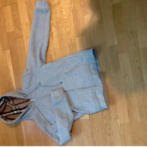 Grå hoodie från Burberry med dragkedja - Säljer en grå hoodie från Burberry med klassiskt rutigt foder i huvan och broderad logga på bröstet. Tröjan har snörning i huvan, två fickor fram och hel dragkedja. Perfekt för dig som gillar stilren design med lyxig touch.