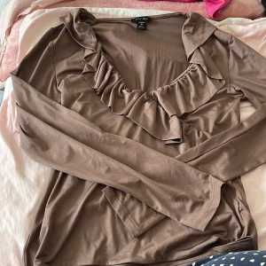 Brun volangblus från Amisu XL - Säljer en brun långärmad blus från Amisu i storlek XL. Toppen har snygg v-ringning med volangdetaljer längs halsringningen och är tillverkad i ett mjukt, lätt material. Perfekt för dig som gillar en feminin och trendig look.