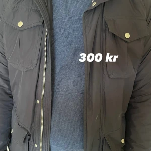 Svart parkajacka från J.Lindeberg - Svart parkajacka från J.Lindeberg med flera fickor och snygga guldfärgade knappar. Jackan har lång ärm, dragkedja och patch på armen. Materialet är polyester och jackan har en normal passform. Perfekt för kalla dagar och har en cool urban vibe. Har två defekter vid varsin ficka som går att fixa! 