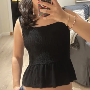 Svart smockad linnetopp med volang - Säljer en svart linnetopp med smockad överdel och breda axelband. Toppen har en söt volangkant nedtill. Perfekt att styla med jeans eller kjol för en chill och trendig look.
