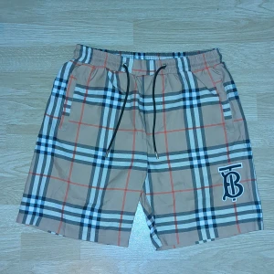 En Äkta Beige rutiga Burberry shorts  - Snygga beige shorts från Burberry med klassiskt rutigt mönster i svart, vitt och rött. De har elastisk midja med svart snörning och Burberrys TB-logga på ena benet. Perfekta för sommaren och har fickor både fram och bak. Materialet är mjukt och lätt polyester.