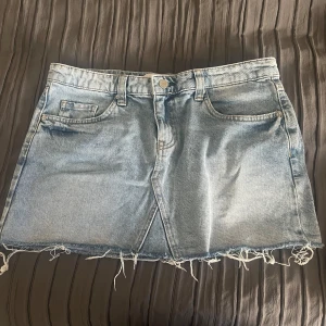Ljusblå jeanskjol från Bershka - Säljer en ljusblå jeanskjol från Bershka med råa, fransiga kanter och klassisk femficksdesign. Kjolen är kort och har en avslappnad vibe, perfekt till sneakers eller boots. Materialet är denim och modellen har bälteshällor och knappstängning framtill.