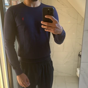 Mörkblå långärmad tröja från Ralph Lauren - Snygg mörkblå långärmad tröja från Ralph Lauren med röd broderad logga på bröstet. Tröjan har rund hals och ribbade muddar vid ärmslut och midja. Perfekt för dig som gillar stilrena och klassiska plagg med en sportig touch. 