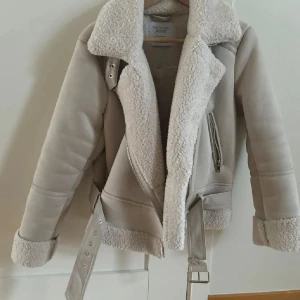 Beige teddyjacka från Stradivarius - Säljer en beige teddyjacka från Stradivarius med fluffigt foder och bred krage. Jackan har coola detaljer som bälte i midjan, dragkedja och muddar med teddy. Perfekt för dig som vill ha en varm och snygg look under hösten och vintern.