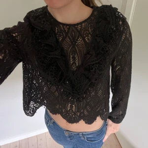 Svart spetsblus med volangdetaljer - Snygg svart blus i spets med lång ärm och croppad passform. 