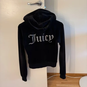 Svart Juicy Couture hoodie med strass - Ikonisk svart hoodie från Juicy Couture i mjuk velour med strassdetaljer. Stor Juicy-text i strass på ryggen och ett glittrande J på bröstet. Dragkedja framtill med J-berlock, huva och känguruficka. Perfekt för dig som gillar Y2K-vibe och comfy streetstyle.