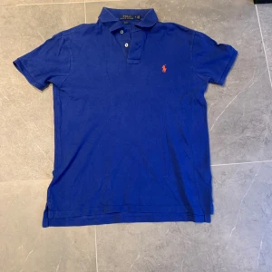 Polo Ralph Lauren Slim Fit Piké – Blå (M) - Säljer en klassisk Polo Ralph Lauren pikétröja i färgen blå med den ikoniska röda loggan på bröstet. 	•	Modell: Slim Fit 	•	Storlek: M 	•	Färg: Blå med orange logga 	•	Skick: Bra begagnat skick, inga hål eller fläckar. 	•	Nypris: ca 1 199 kr  Perfekt för både vardag och lite mer uppklädda tillfällen.  💰 Mitt pris: 250 kr