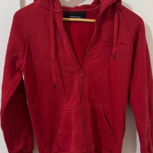 Röd Peak Performance hoodie med dragkedja - Säljer en röd hoodie från Peak Performance i storlek S. Tröjan har huva med snörning, broderad logga på bröstet, två fickor fram och hel dragkedja. Materialet är mjuk bomull, perfekt för chill eller träning. Långärmad och med ribbade muddar.