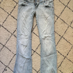 Ljusblå bootcut jeans från Lee W27 L33 - Säljer ett par ljusblå jeans från Lee med bootcut passform och klassisk femficksdesign. Jeansen har slitningar vid ena knät, orangea sömmar och dragkedja i gylfen. 