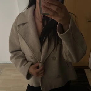 Beige kavajjacka med bred krage - Snygg beige kavajjacka med bred krage och dubbelknäppning framtill. Jackan har en loose passform och är tillverkad i ett mjukt ullmaterial som ger en lyxig känsla. Perfekt för dig som gillar stilrena och trendiga plagg med lite oversized look.