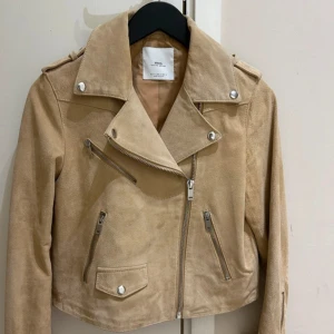 Beige skinnjacka från MNG - Snygg beige skinnjacka från MNG med coola silvriga detaljer och flera dragkedjor framtill. Jackan har klassisk bikerstil med bred krage och axelklaffar. Croppad passform som ger en trendig look, perfekt till jeans eller klänning.