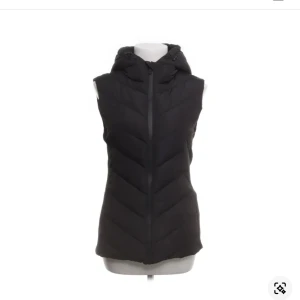 Svart dunväst med huva från Zara - Svart dunväst från Zara med snyggt chevronmönster och praktisk huva. Västen har dragkedja framtill och är lätt vadderad för extra värme. Perfekt att slänga över en hoodie eller tröja när det är kyligt ute.