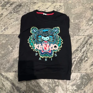 Svart Kenzo sweatshirt med tigertryck - Svart sweatshirt från Kenzo i storlek M med ikoniskt broderat tigerhuvud i blått, grönt och gult framtill. Tröjan har rund hals, långa ärmar och ribbade muddar. Kenzo Paris-loggan är broderad över motivet. Perfekt statement-plagg för dig som gillar streetstyle.