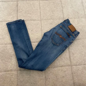 Blå slim jeans från Nudie Jeans - Säljer ett par blå slim jeans från Nudie Jeans med klassisk femficksdesign och snygga kontrastsömmar på bakfickorna. Jeansen har normal passform i midjan och är tillverkade i mjukt jeansmaterial med lätt tvättad look. Perfekt för dig som gillar stilrena och bekväma jeans.