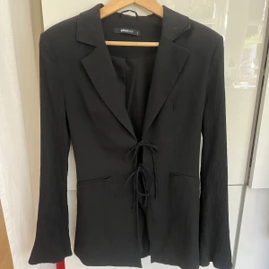 Svart kavaj med snörning från Gina Tricot - Svart kavaj från Gina Tricot med snygg snörning framtill istället för knappar. Modellen har lätt utsvängda ärmar och är gjord i ett strukturerat, krinklat tyg som ger cool textur. Klassisk krage och två fickor framtill. Perfekt för dig som gillar stilrena detaljer. Kavajen är knappt använd endast ett fåtal gånger. Säljer pga den inte används. Väldigt bra skick.