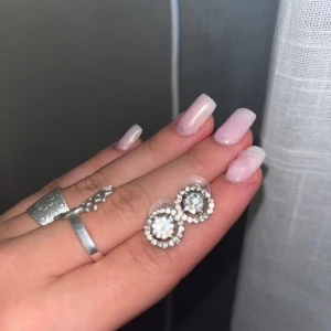 Glittriga silverörhängen med stenar - Nypris 300kr. Säljer ett par snygga örhängen i silverfärg med rund form och glittriga stenar runt en större kristall i mitten. Perfekta om du vill addera lite extra bling till din look. De är små och diskreta men ändå riktigt iögonfallande. 