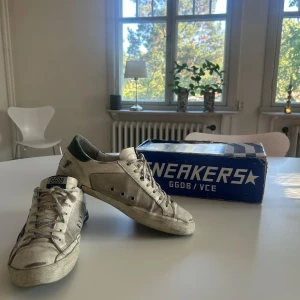 Golden Goose vita sneakers med stjärna - Golden Goose sneakers i vit canvas med svart stjärna på sidan och gröna detaljer vid hälen. Skorna har snörning med GGDB-tryck på skosnörena och en platt sula i gummi. Snyggt slitna look och klassisk rund tå, perfekt för dig som gillar streetstyle. 