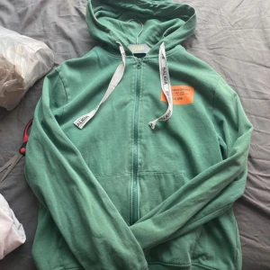 Grön hoodie med dragkedja från Saligia - Grön hoodie från Saligia i storlek medium med vit snörning där det står 'SALIGIA'. Tröjan har dragkedja framtill, huva och en orange patch med text på bröstet. Perfekt för chill dagar och streetstyle.