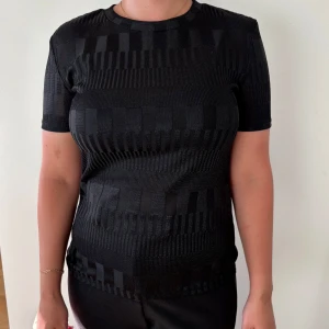 Svart mönstrad t-shirt - Svart t-shirt med diskret geometriskt mönster i olika nyanser av svart. Modellen är kortärmad och har rund hals. Materialet är stickat och ger en cool struktur till plagget. 