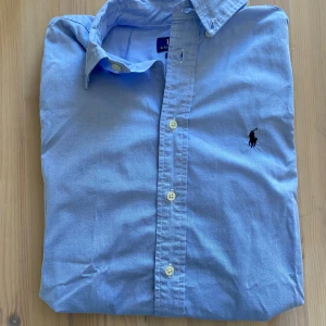 Ljusblå skjorta från Polo Ralph Lauren - Klassisk ljusblå skjorta från Polo Ralph Lauren med broderad logga på bröstet. Skjortan har slim fit-passform, långa ärmar och knäppning framtill. Perfekt för en stilren look.