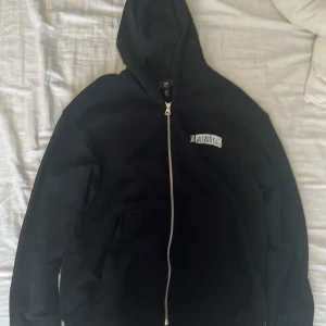 Svart hoodie med dragkedja från H&M - Svart hoodie från H&M med dragkedja och huva. Broderad text 'LA PRIVILEGES' på bröstet och ryggen. Modellen är loose fit och har klassisk design med fickor framtill. Perfekt för en avslappnad stil.